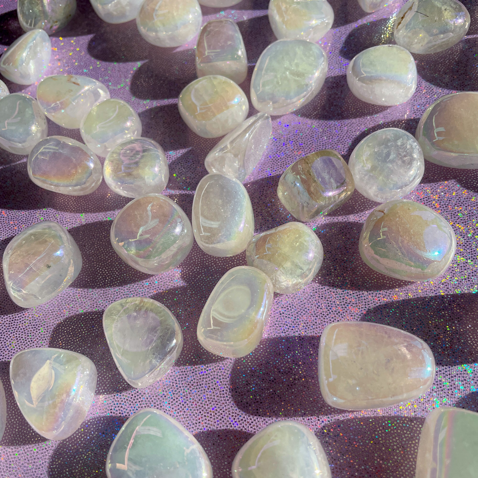 Small-sized, smooth, clear-ish white with iridescence, Angel Aura Quartz tumbled pocket stones against a sparkly purple background.