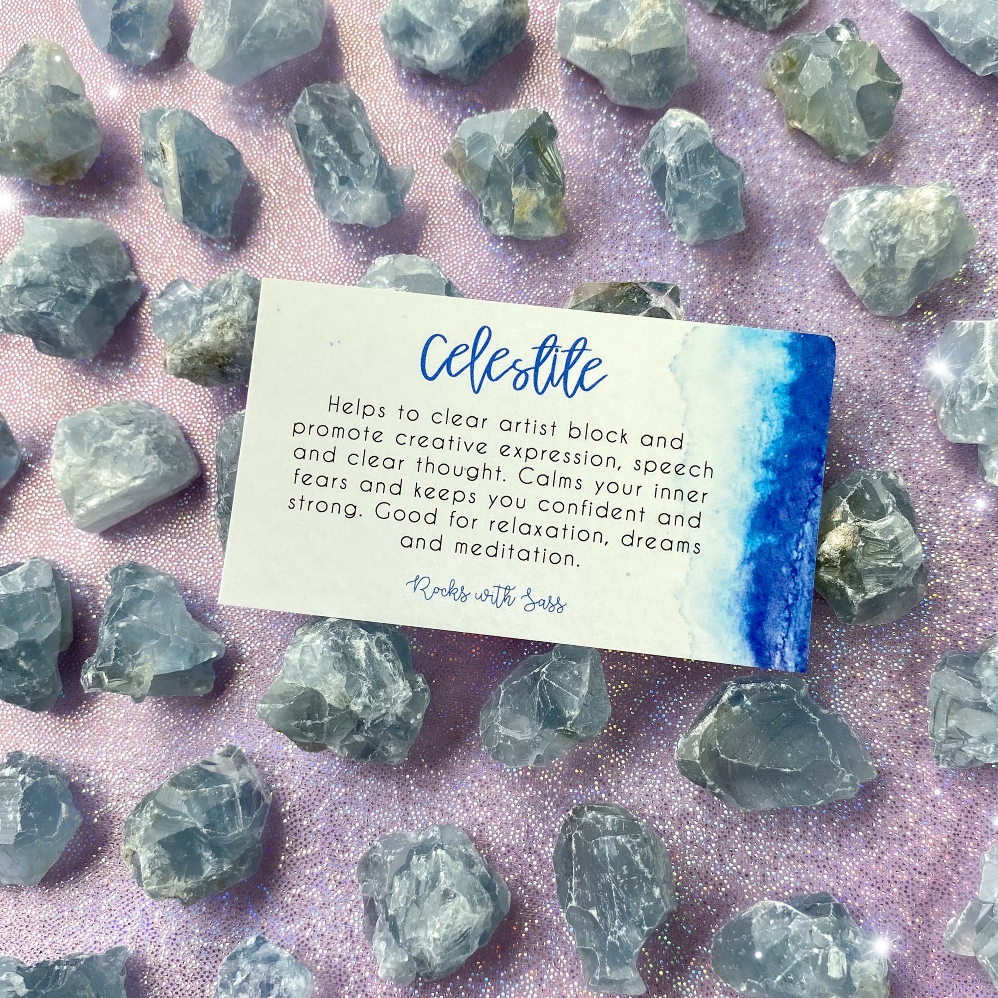 Small-sized, rough, blue, raw Celestite pocket stones against a purple sparkly background with a "Celestite" meaning card laying on top of them.