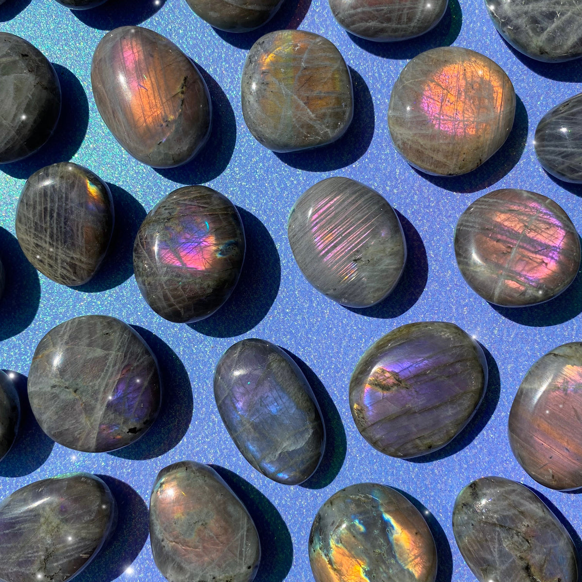 Vivid, flashy, vibrant-colored, Sunset Labradorite palm stones against a blue, sparkly background.