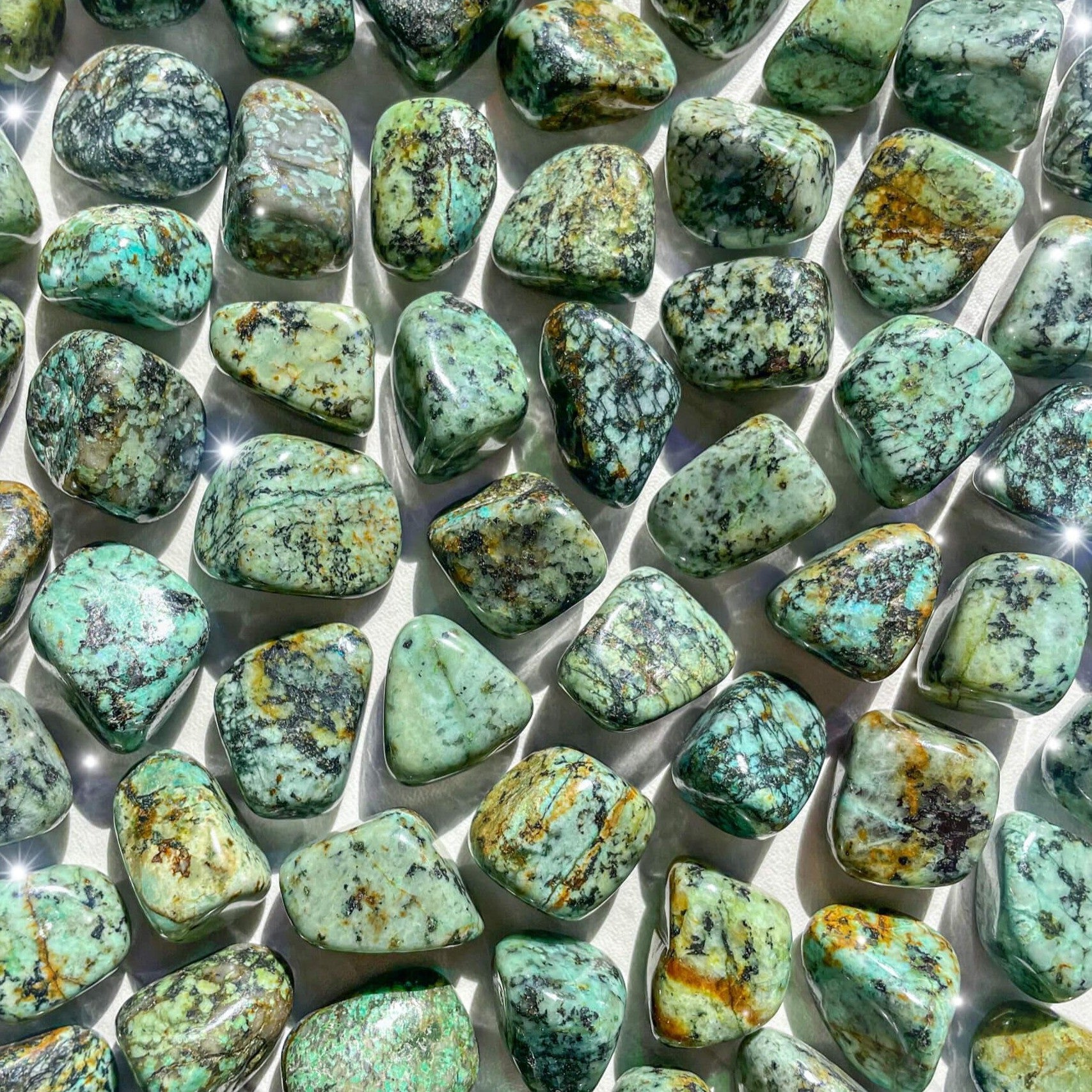 Small-sized, smooth, blue, green, and brown African Turquoise tumbled pocket stones against a white background.