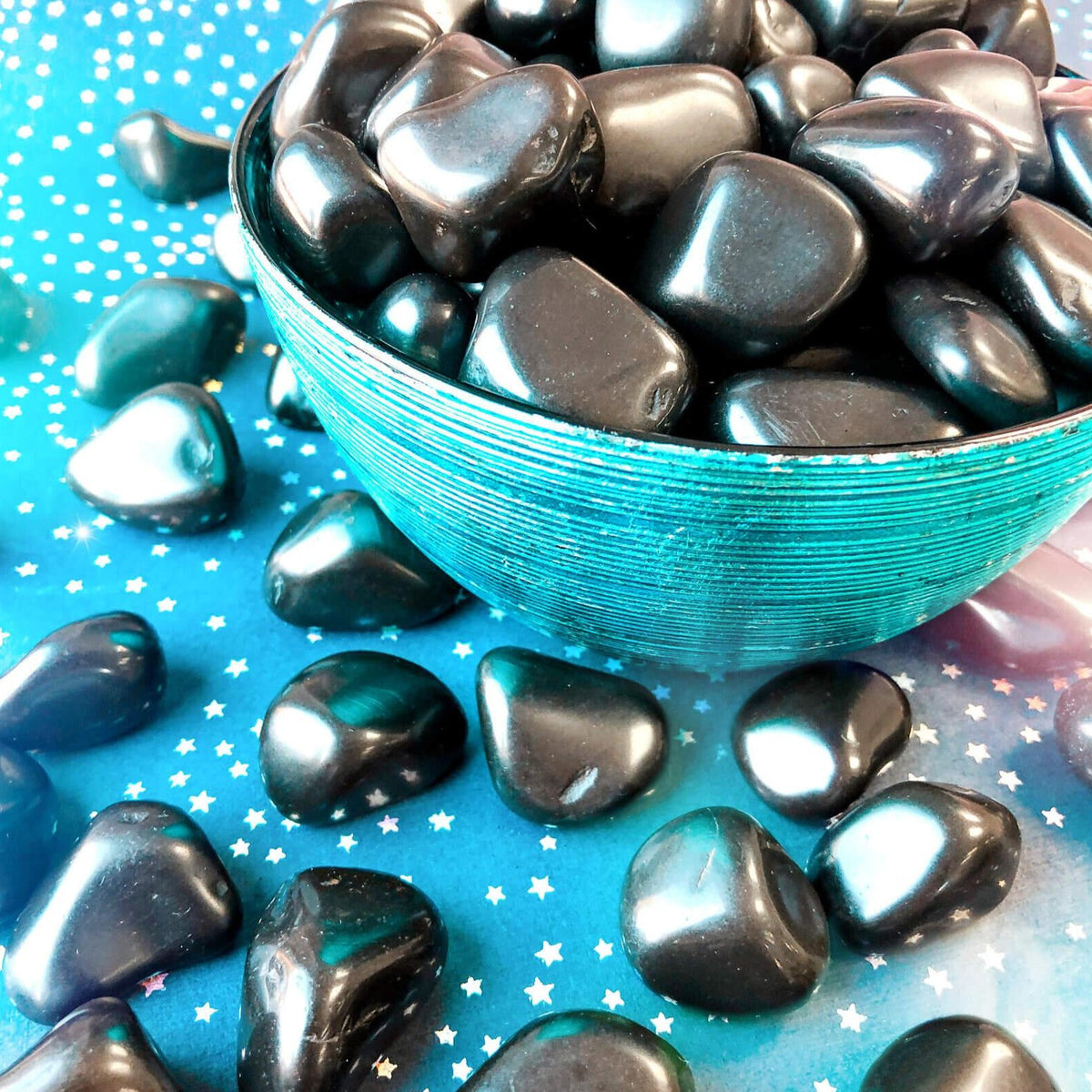 Small-sized, smooth Black Tourmaline tumbled pocket stones in a blue bowl against a blue, starry background.