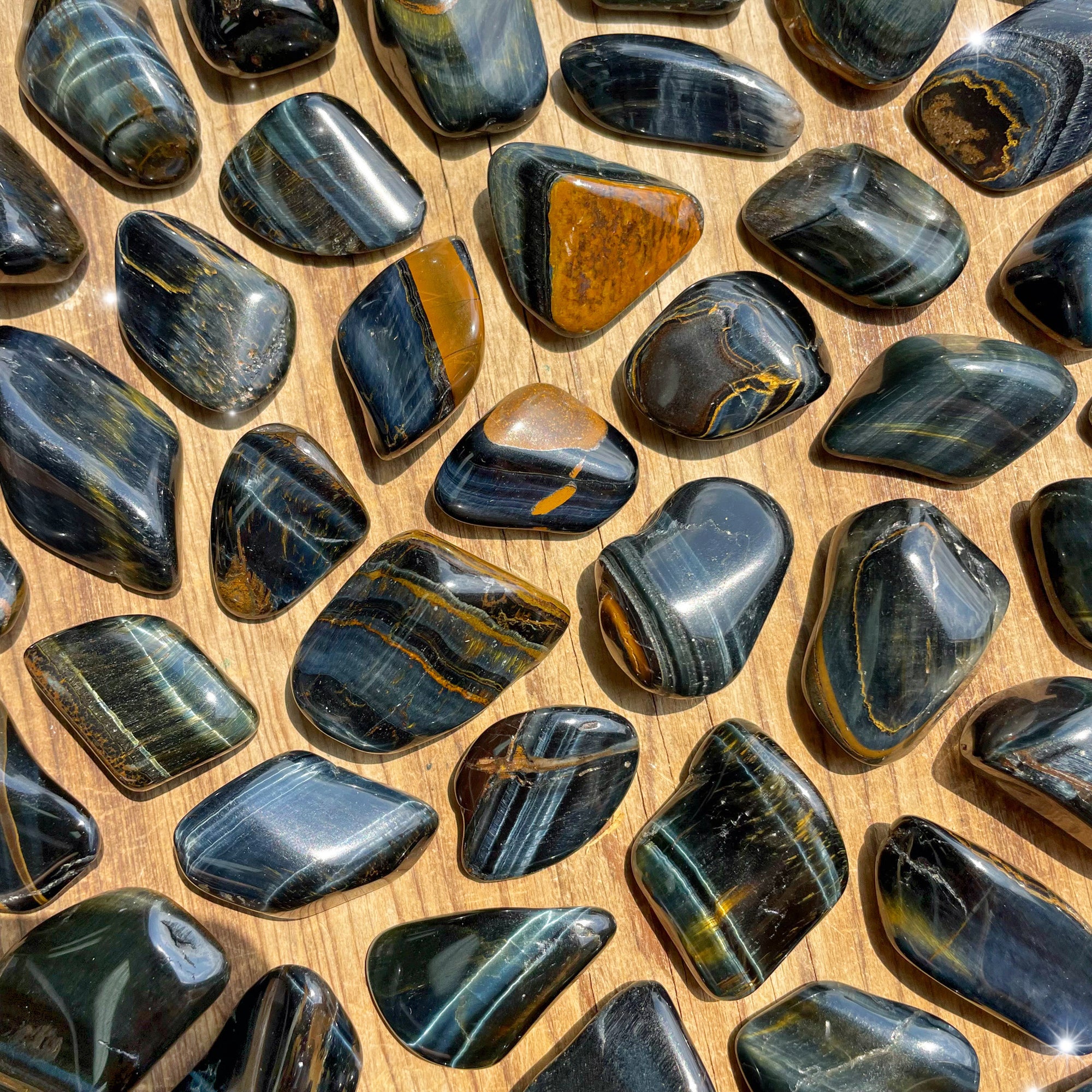 Small-sized, smooth, blue and brown striped, shiny, Blue Tiger's Eye tumbled pocket stones laying against a wooden background.